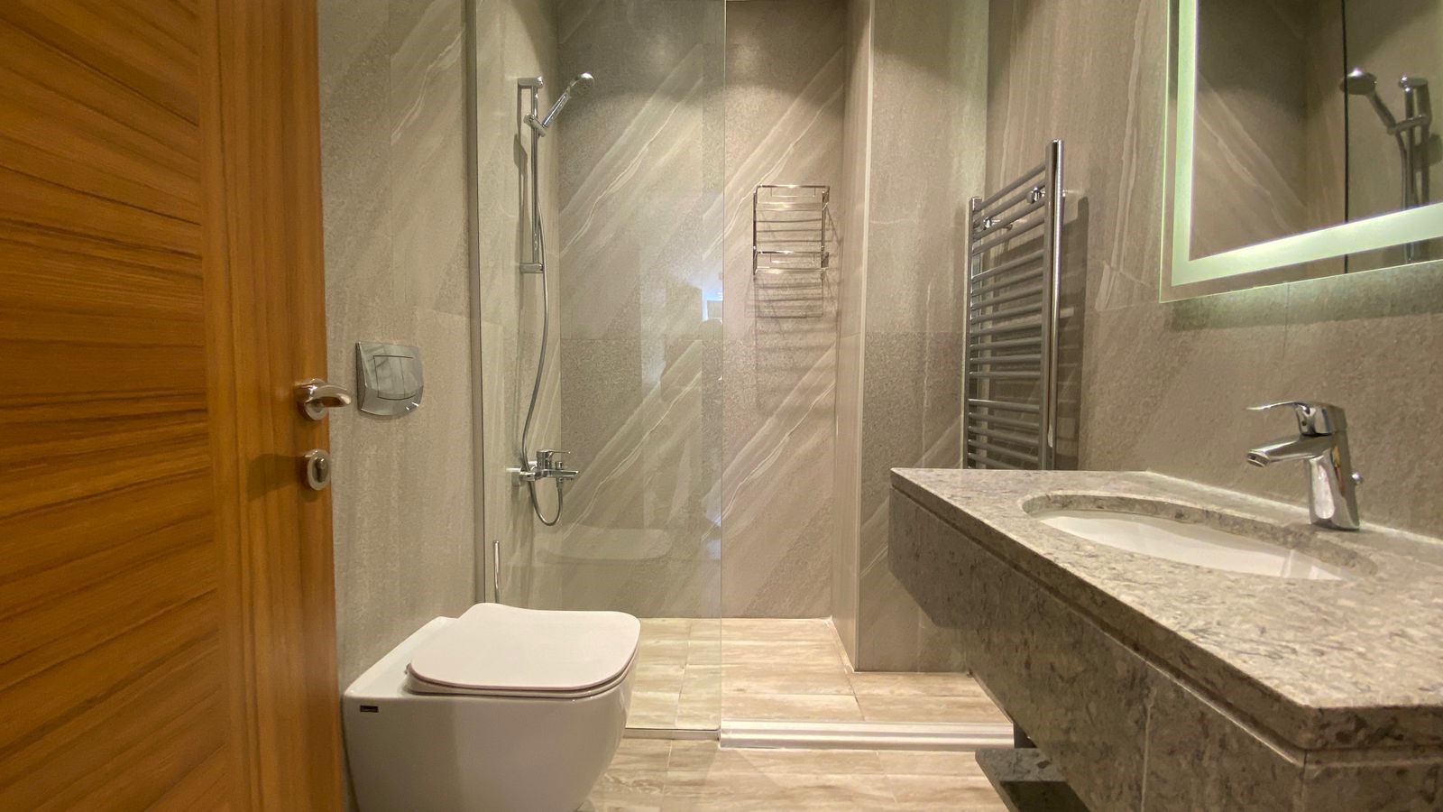 3d615105-f24c-44b0-9bc6-bea60720a63e Modern bathroom with a wooden door on the left, a wall-mounted toilet, and a glass shower enclosure beside a granite countertop sink.