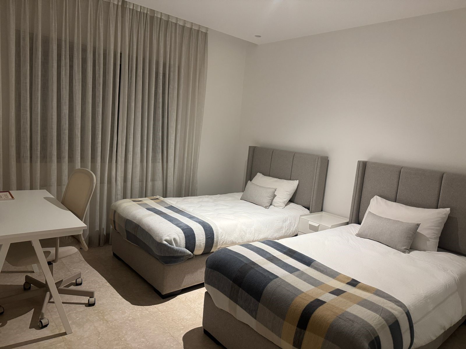 3e73ad48-b11d-4321-8390-3471a95ee59f Twin hotel room with two gray upholstered beds, white bedding, and a striped throw; desk and chair by a large window with sheer curtains.