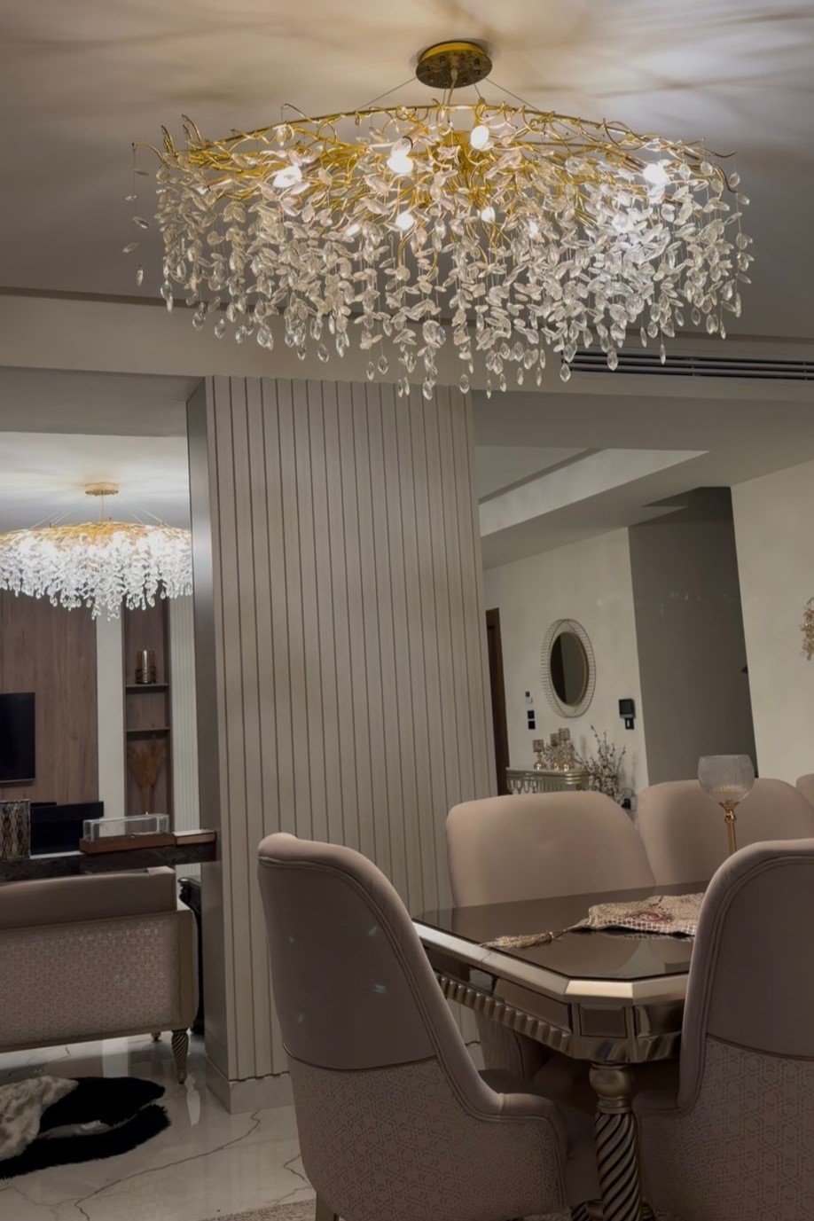 3fc9a0f7-fbeb-4002-86fb-5decd0240d3f Elegant dining room with a crystal leaf chandelier above a glass-topped table and beige upholstered chairs.