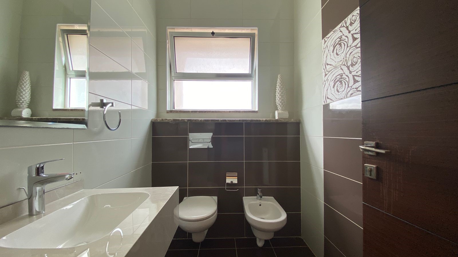 40a70409-97a8-42f5-be21-faade53b7412 Modern bathroom with a white sink, chrome faucet, toilet and bidet opposite a dark tiled wall; window above and decorative white sculptures on the ledge.