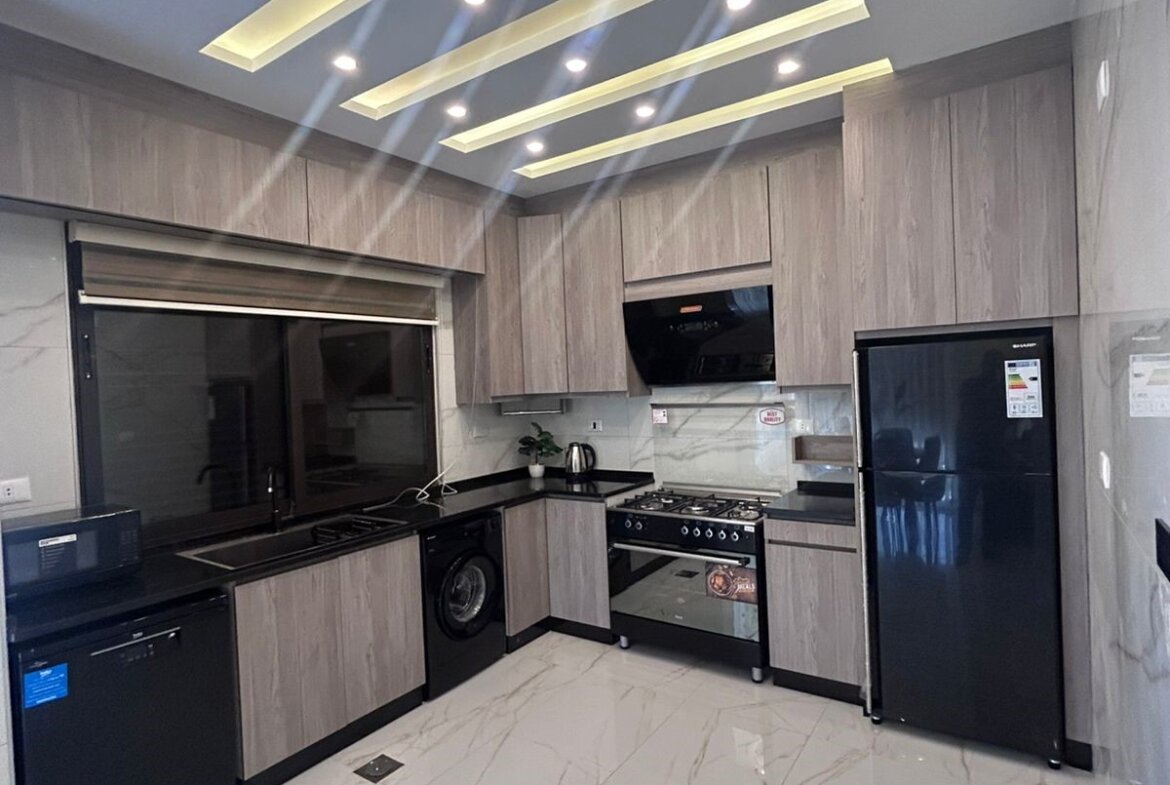 Modern L-shaped kitchen with grey wood cabinets, black countertops, a stainless fridge, oven, dishwasher, and washing machine under the counter across white marble flooring.
