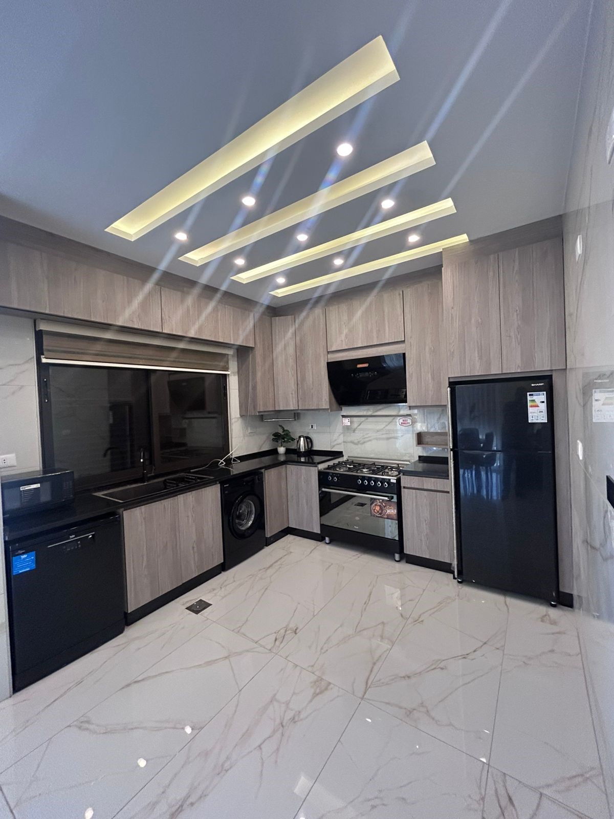 40d86f1d-3474-4e67-8716-8b7a922d0010 Modern L-shaped kitchen with grey wood cabinets, black countertops, a stainless fridge, oven, dishwasher, and washing machine under the counter across white marble flooring.