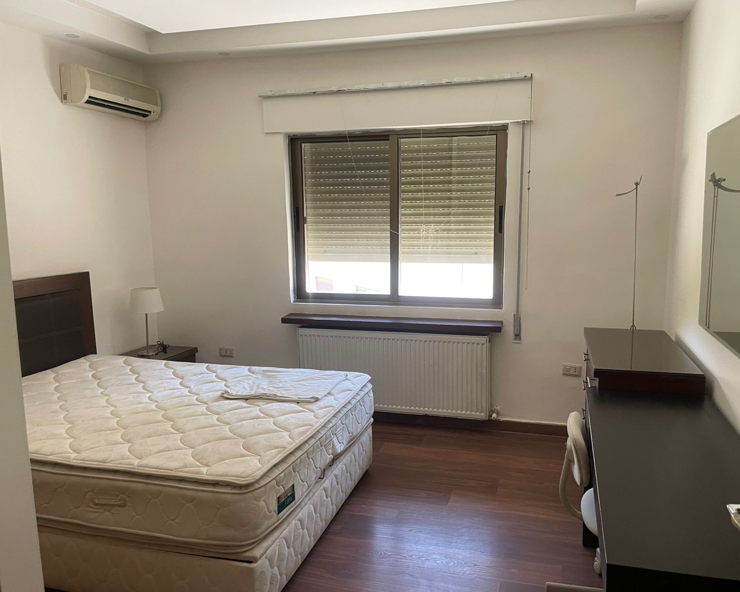 41af5c2e-8518-4112-bb11-d546328acf90 Small bedroom with a double bed on a dark wood frame, mattress exposed, a bedside lamp on a nightstand, window with partially closed blinds, radiator beneath the window, and a dresser with a mirror nearby.