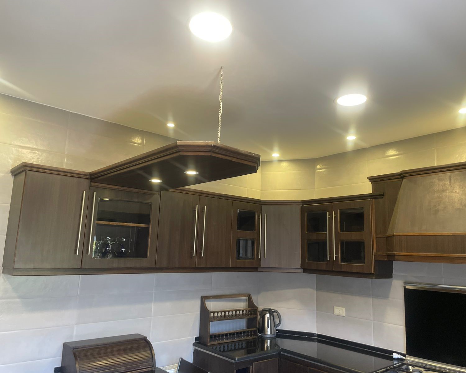 429b3518-1160-470b-a75c-a3a3bbb595d4 Dark wood kitchen cabinets with glass-front doors and a suspended wooden range hood above the stove, chain visible against a light ceiling.