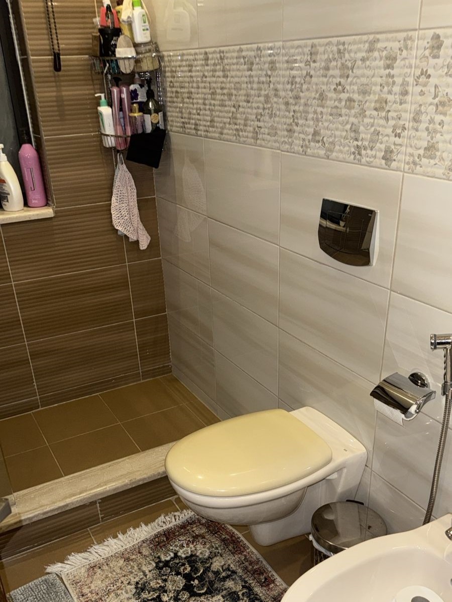 42cfd120-f2db-4257-ae95-81163f836730 Bathroom with a wall-hung toilet, a bidet sprayer, and a chrome flush plate on white tiled wall; brown tiled shower area to the left with shelves of toiletries; decorative rug on the floor.