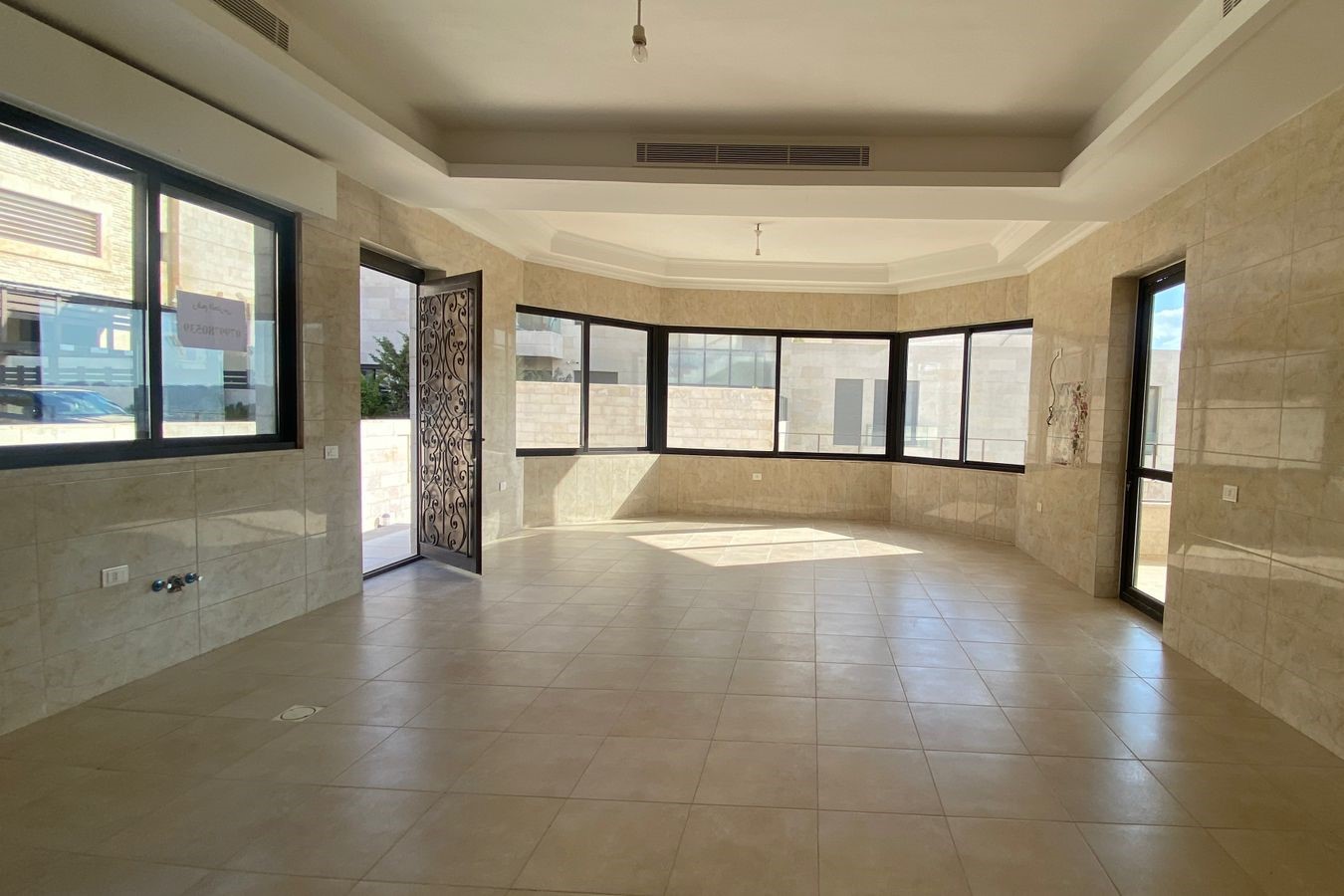 43d11c4c-66f1-4ba6-83e7-28f8d5df9219 Spacious, empty sunlit room with beige tiled floor and large black-framed windows along a curved wall.