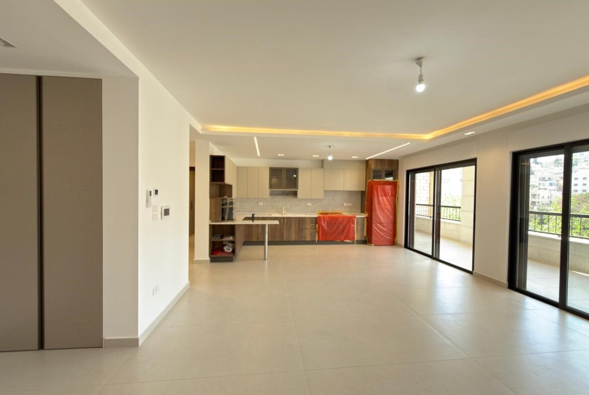Open-plan living area with a modern kitchen, large glass doors to a balcony, and red protective covers on appliances; space appears under setup or construction.