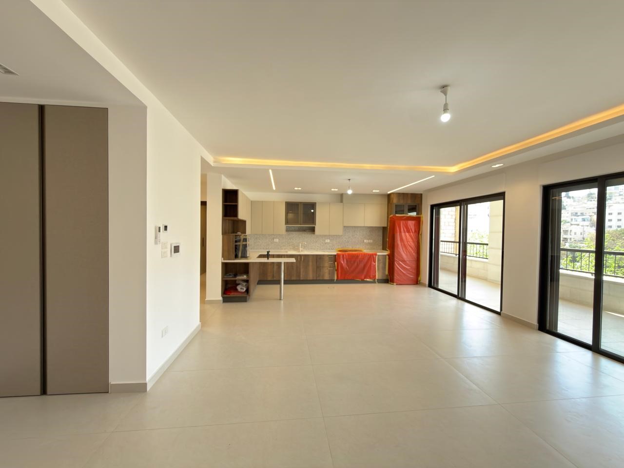 44e7b0a9-e149-4b22-8a4a-3141993045e0 Open-plan living area with a modern kitchen, large glass doors to a balcony, and red protective covers on appliances; space appears under setup or construction.