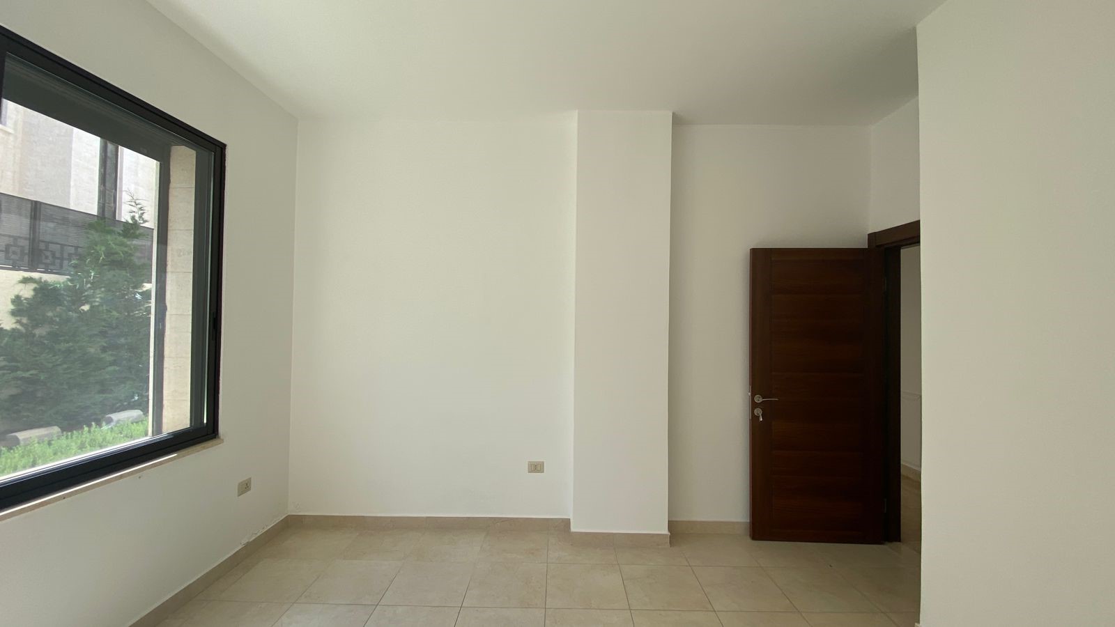 46bfd407-27f1-45a3-834a-9e8e86c907d0 Empty unfurnished room with large left window and an open dark wooden door on the right, beige tiled floor, white walls.