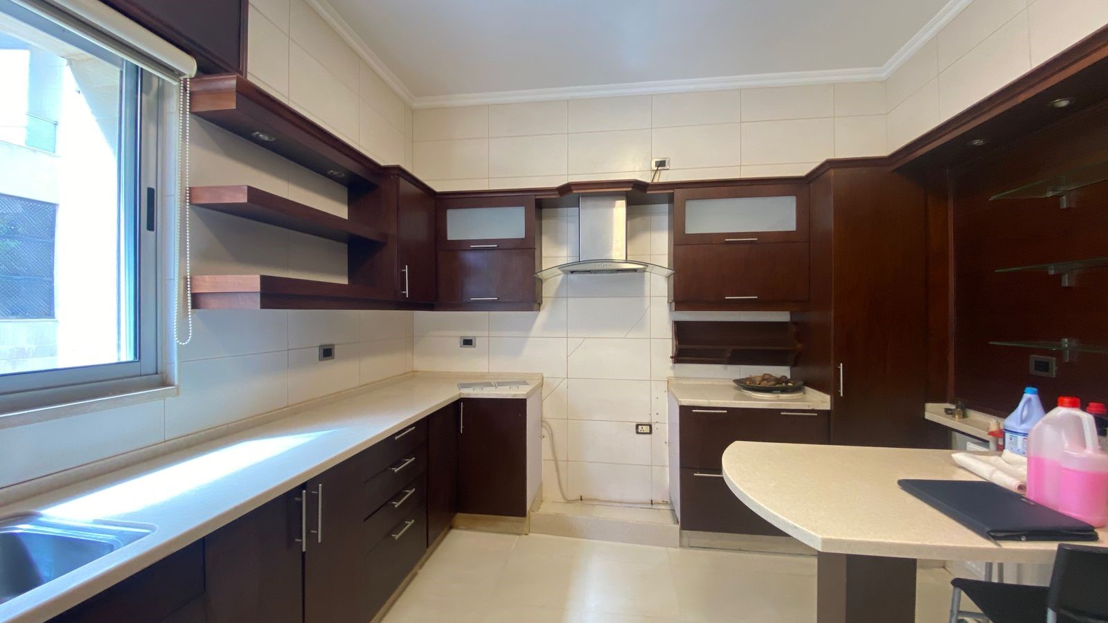 4704533f-1cb3-48d2-bdf0-b0e40eb2df5b U‑shaped kitchen with dark wood cabinets, white counters, and a stainless range hood; window on the left and open shelves nearby.