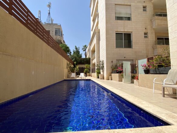 Narrow rectangular blue swimming pool flanked by beige stone walls and a building, with lounge chairs and potted plants along the deck.