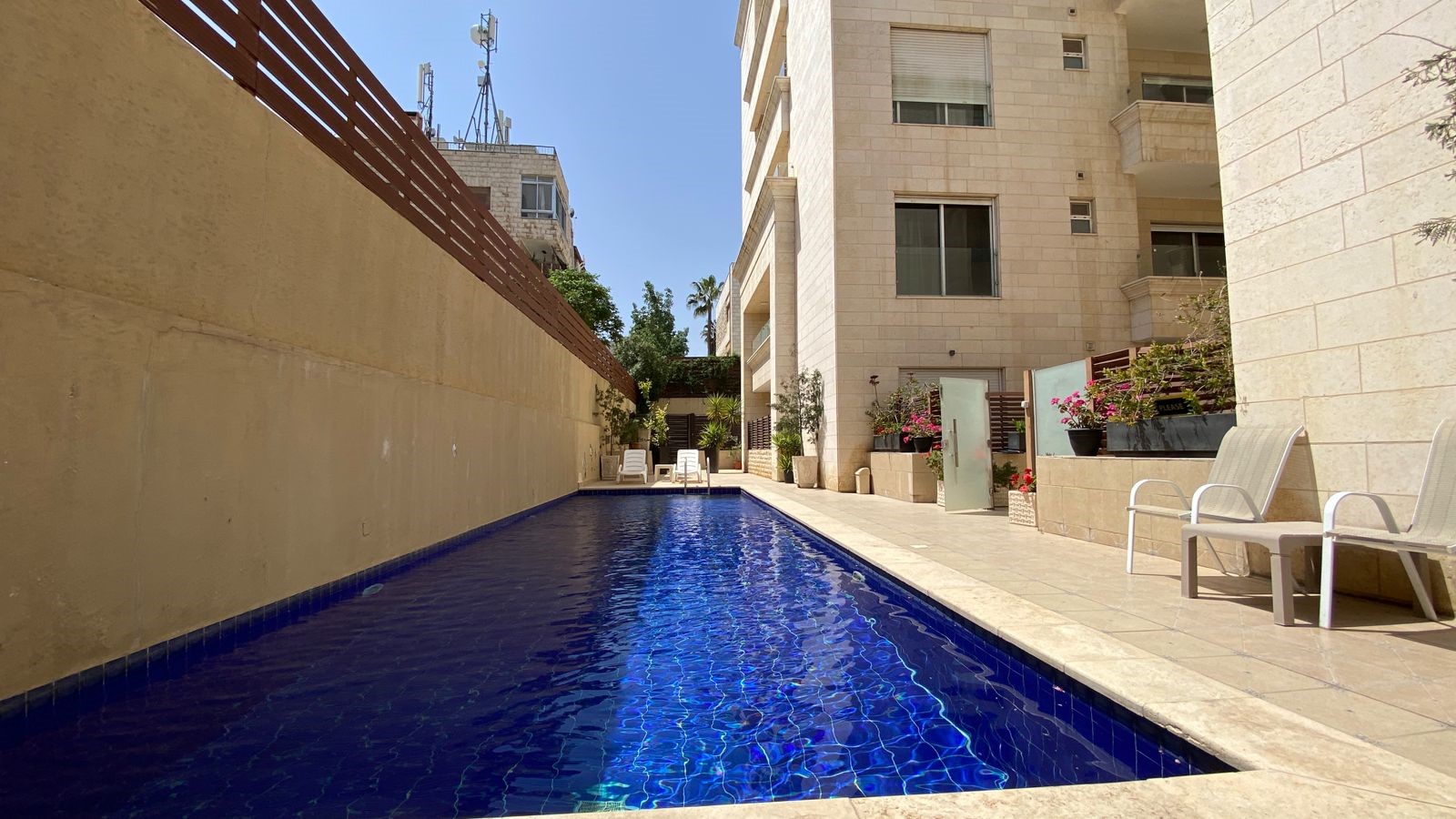 47fd4403-593c-45ad-9806-c48dafd23202 Narrow rectangular blue swimming pool flanked by beige stone walls and a building, with lounge chairs and potted plants along the deck.