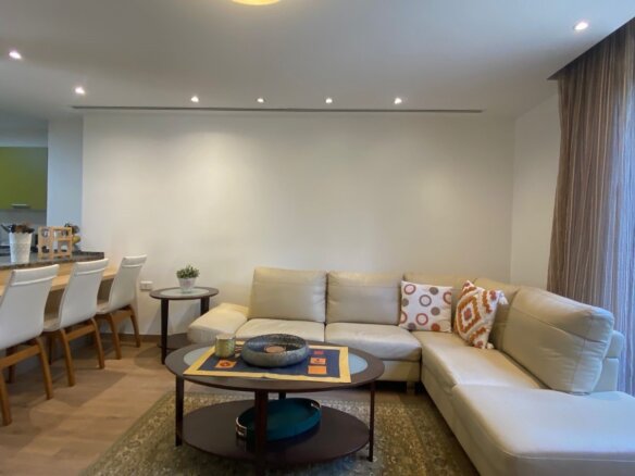 Open-plan living area with a beige sectional sofa, coffee table, and large windows showing greenery outside.
