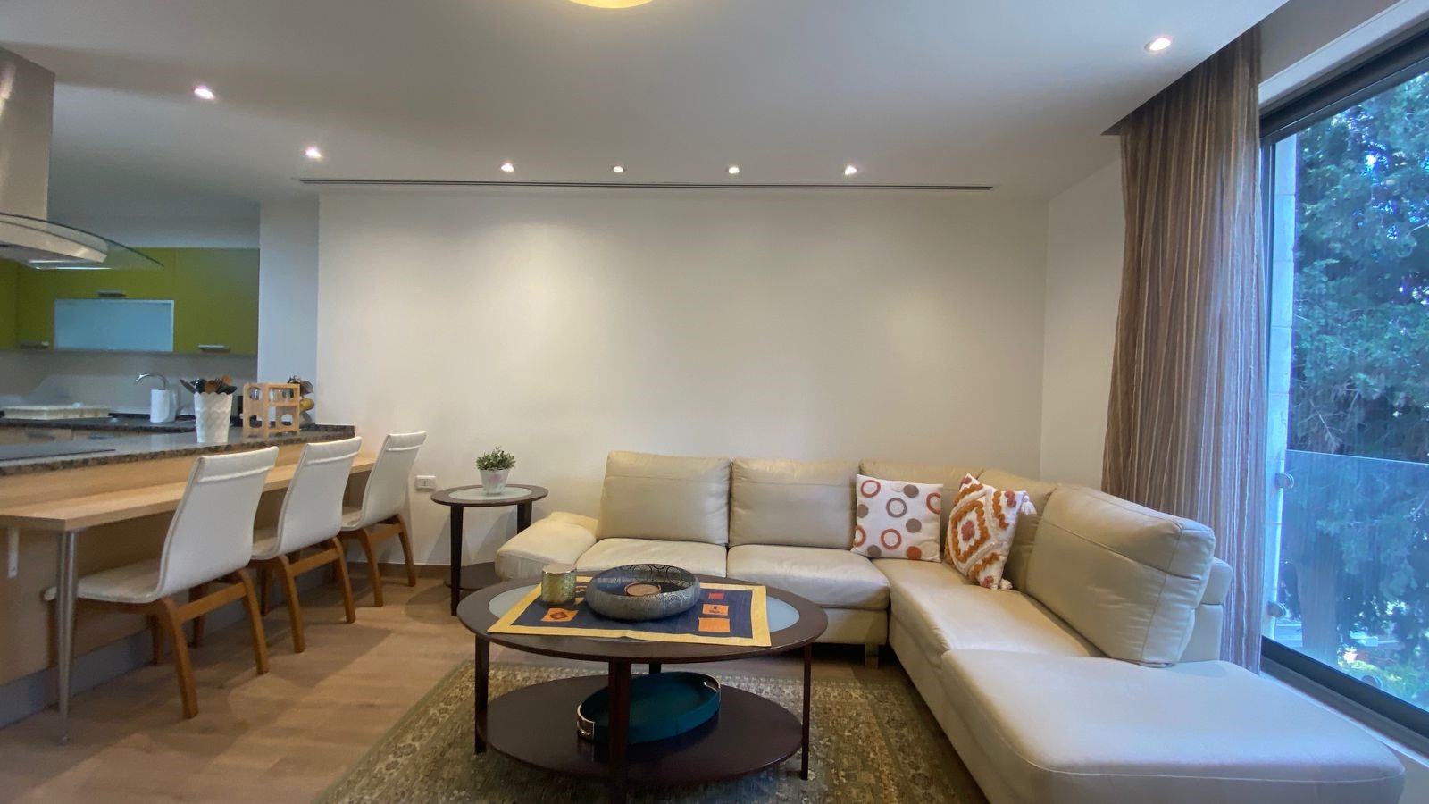 49075f73-35a8-43e5-be9e-2f89877c4a3c Open-plan living area with a beige sectional sofa, coffee table, and large windows showing greenery outside.