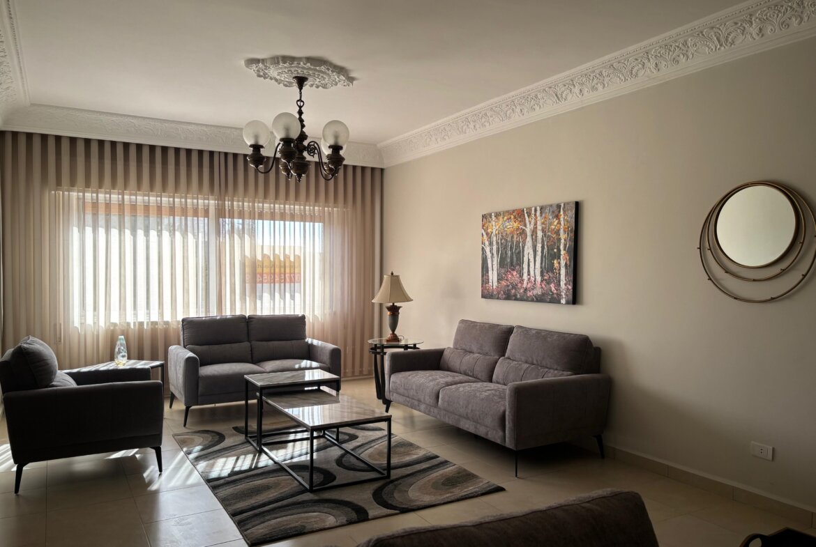 Living room with gray sofas, a glass coffee table, and a patterned rug under natural light from sheer curtains and a chandelier above