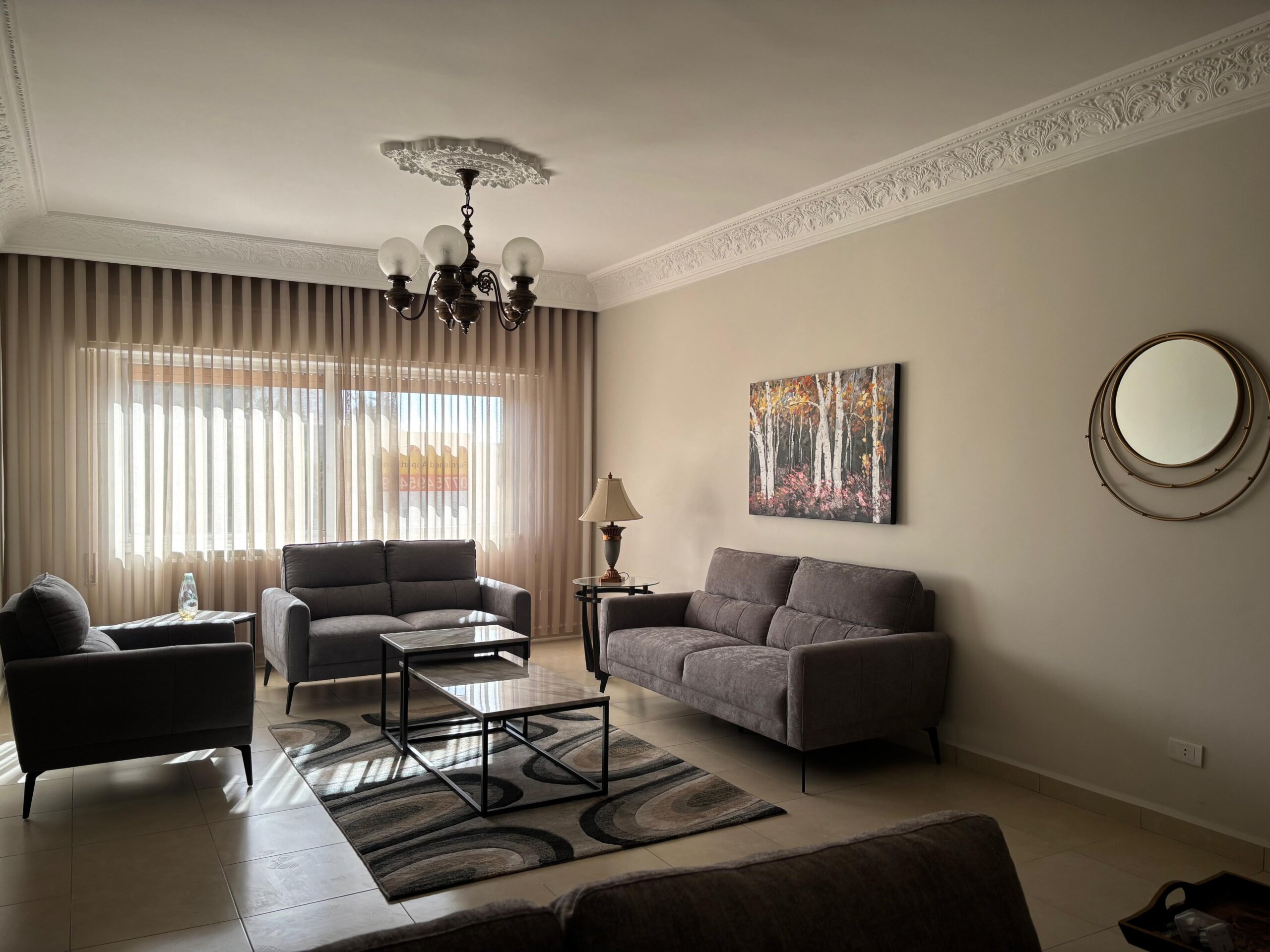 49ad6093-3919-4098-bae3-9a139f0ac8cb Living room with gray sofas, a glass coffee table, and a patterned rug under natural light from sheer curtains and a chandelier above
