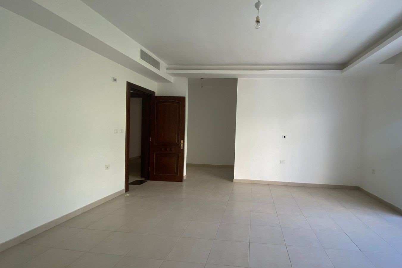 4a801432-7396-4e2d-8423-f30e6a6e674d Empty salon with white walls, beige tile floor, and an open wooden door leading to another room; bare light bulb hanging from ceiling.