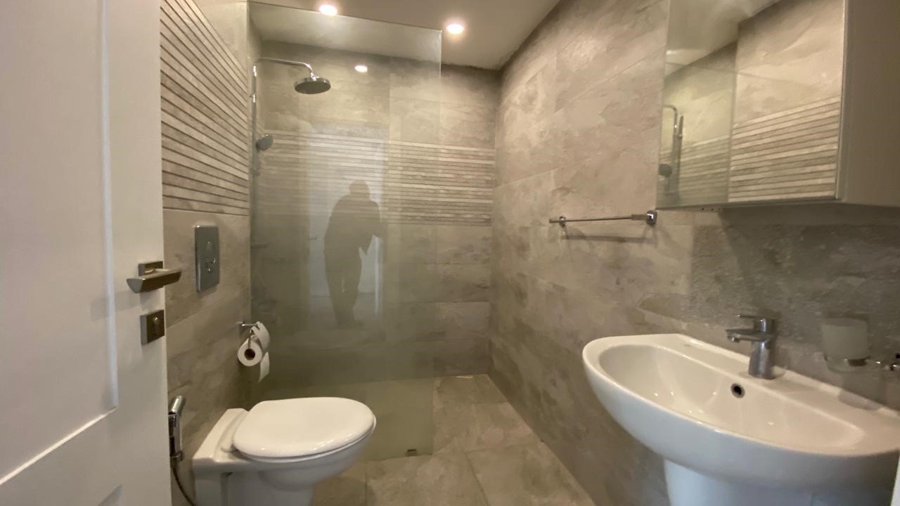 4a8b183f-8602-4042-a11d-000b50f45647 Modern bathroom with glass-walled shower, toilet, and pedestal sink in a marble-tiled room; reflection of a person seen in the glass panel