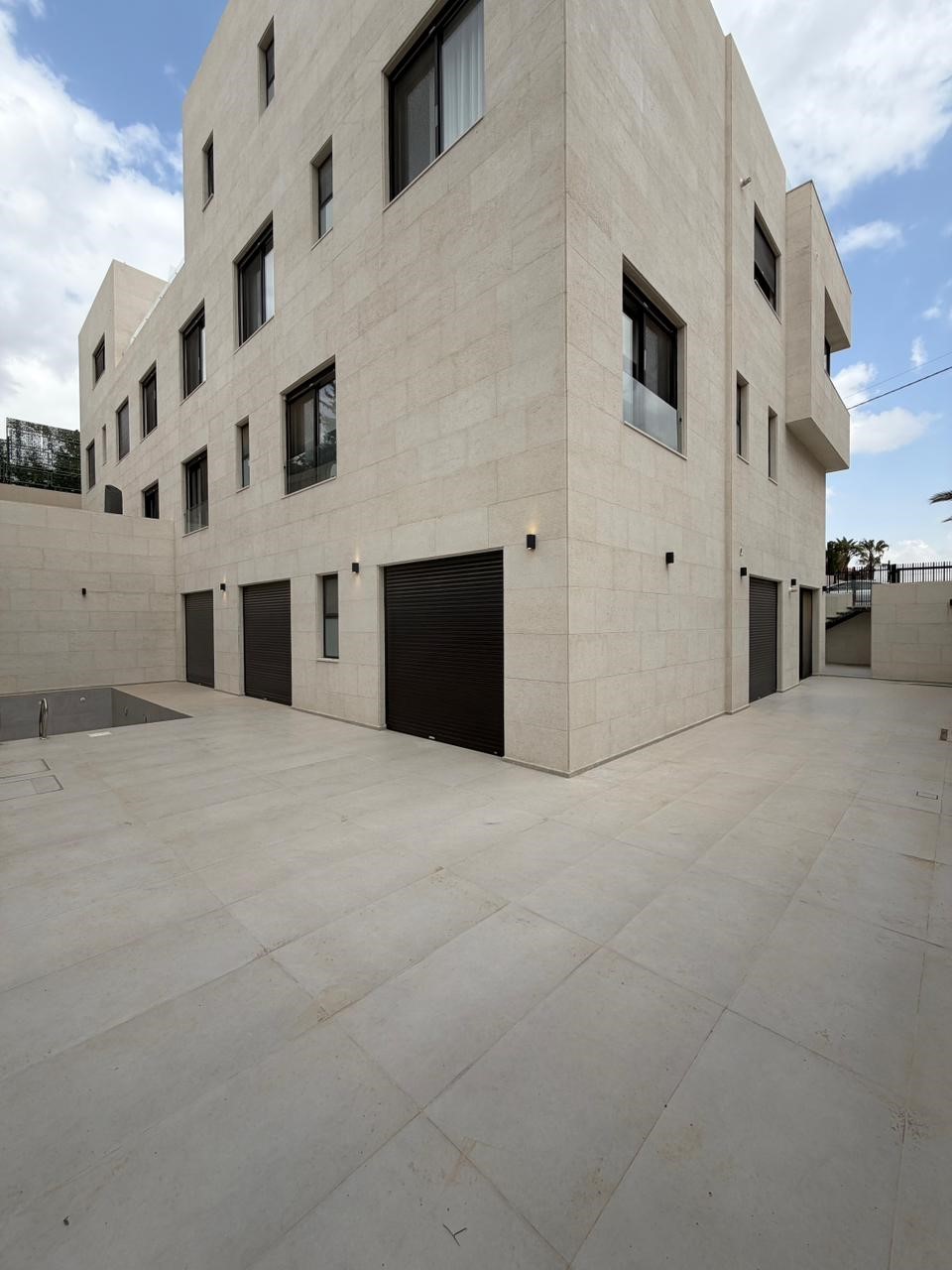4bcf2a71-f7d4-42c7-8264-69da45c35b45 Exterior view of a modern beige concrete building with multiple black garage doors along a wide, tiled courtyard under a blue sky.