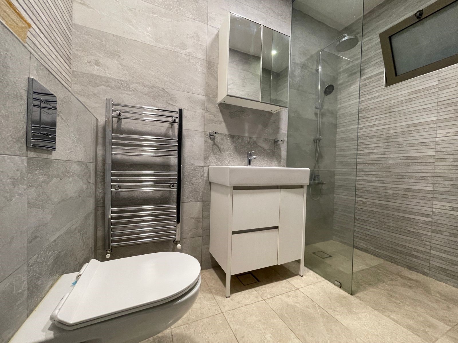 4c2a01fa-ca81-4162-81de-5d3c1d368d47 Modern gray-tiled bathroom with white vanity, wall cabinet, and glass-enclosed shower featuring a chrome towel warmer nearby.