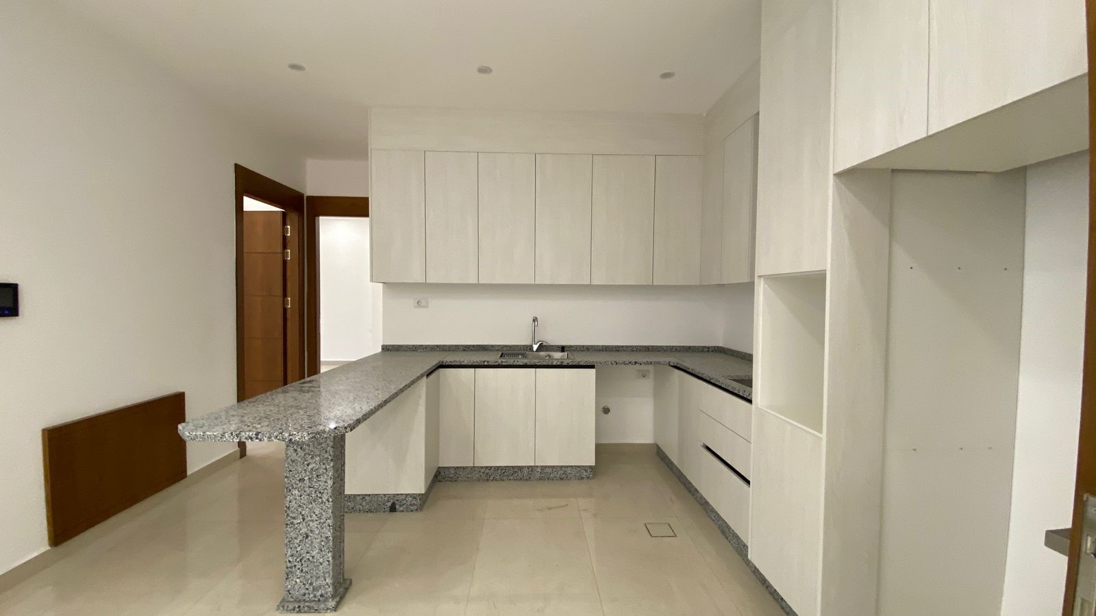 4c38271f-0dd4-460c-9a1b-df6c4c998f76 Modern white kitchen with granite counters and an island, L-shaped cabinets, and a sink in the back wall.
