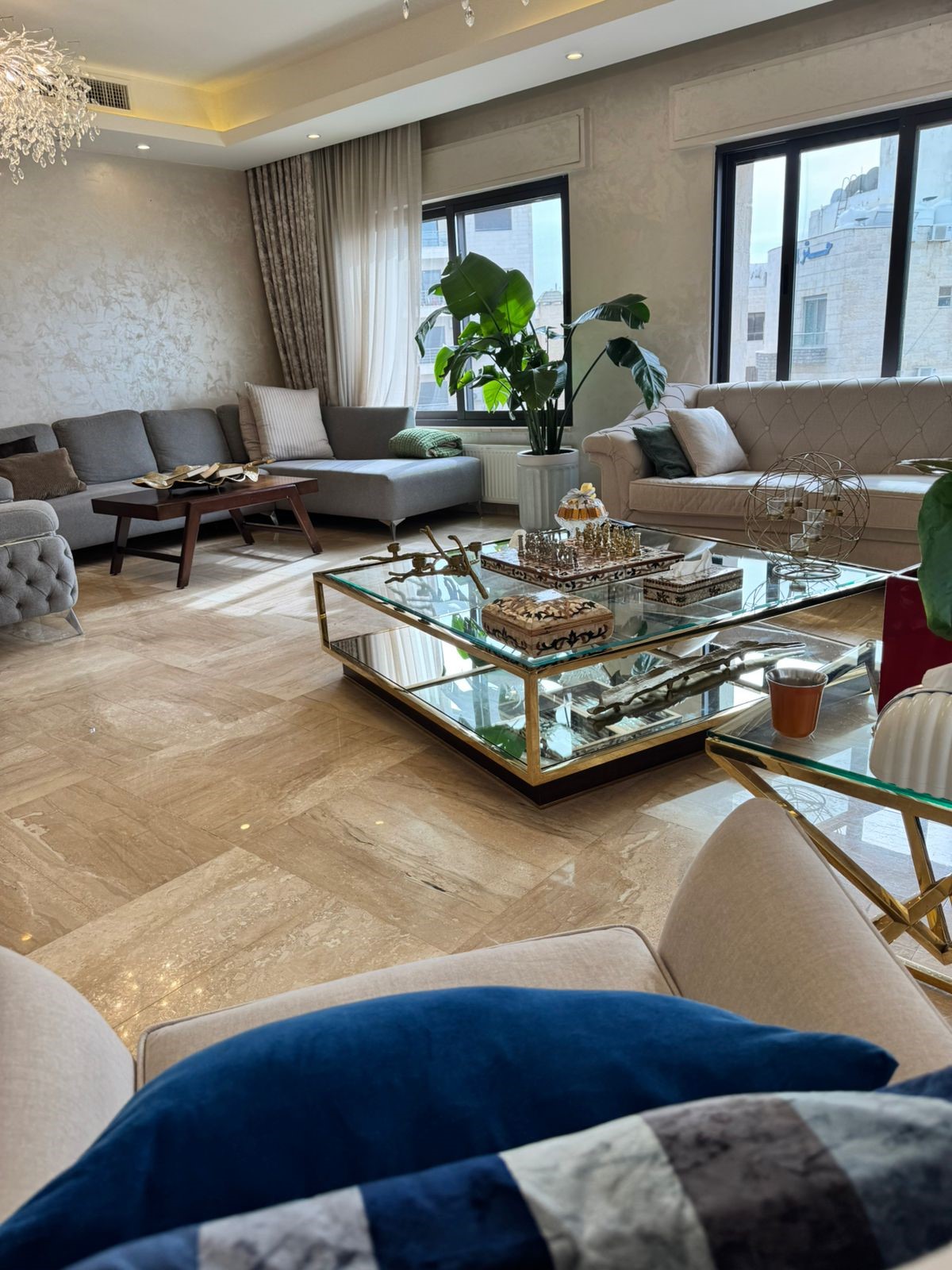 4c4e641f-c59a-431b-b0cc-a30fdec35bc6 Bright living room with beige marble flooring, gray and cream sofas, and a glass coffee table with decorative items.