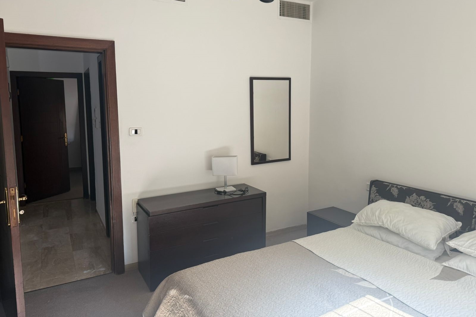 4e0ec111-02ac-4d29-ad4c-83a0b76dd886 Hotel bedroom with a double bed, white bedding, and a dark wood dresser with a lamp on top and a wall mirror nearby.