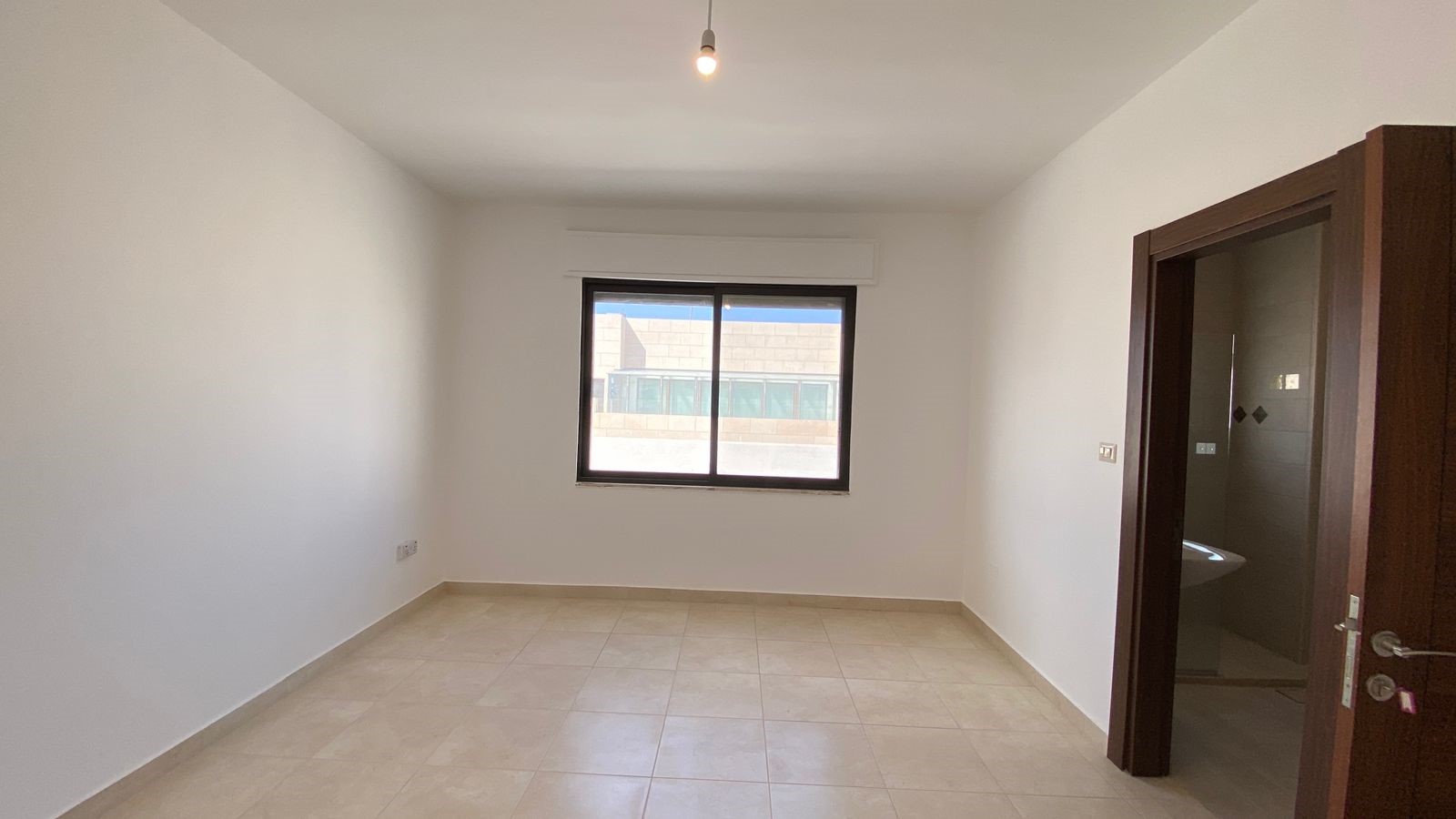 4f5b7153-24fb-4aa4-acd1-b855be65ed1e Empty unfurnished room with beige tiled floor, white walls, a centered window, and an open bathroom door on the right, ceiling bulb visible.