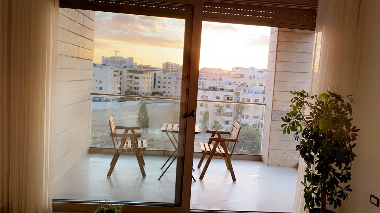 4f77feb5-a90d-42a6-a557-8ccf460735ff Balcony scene at sunset with wooden chairs and a small table, glass railing overlooking a cityscape.