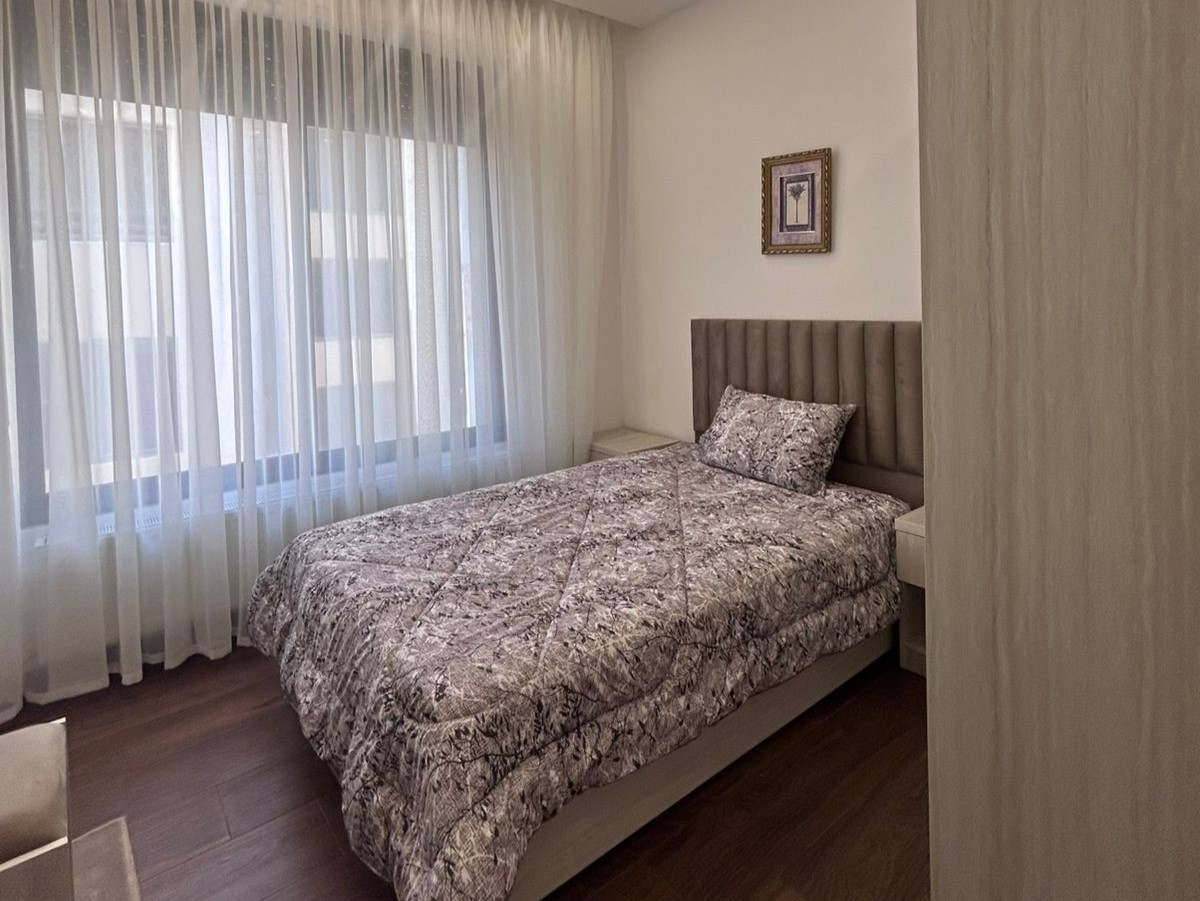 4f8dc3bd-4a2b-435d-baa0-bf0c1d6dd64f Light-filled bedroom with a padded headboard bed, two white nightstands, and sheer curtains along a large window.