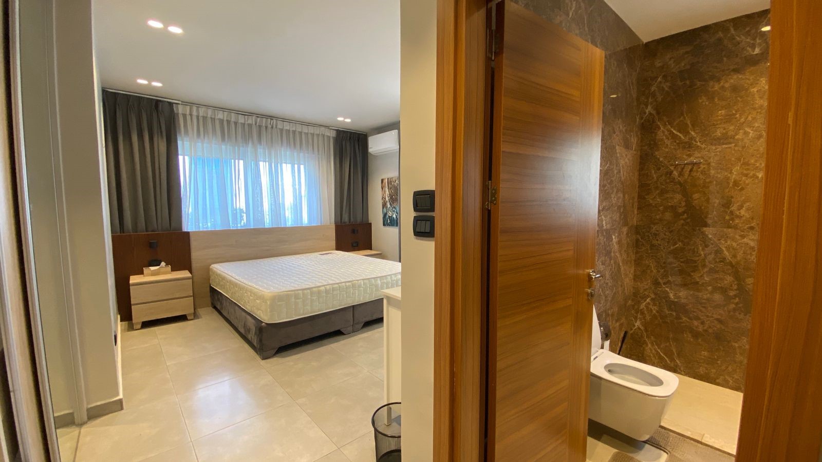 4fba5a73-a646-4764-a56f-5e35e4330b63 Hotel bedroom with a queen bed, two nightstands, and a large window with sheer curtains; marble bathroom visible through an open door.