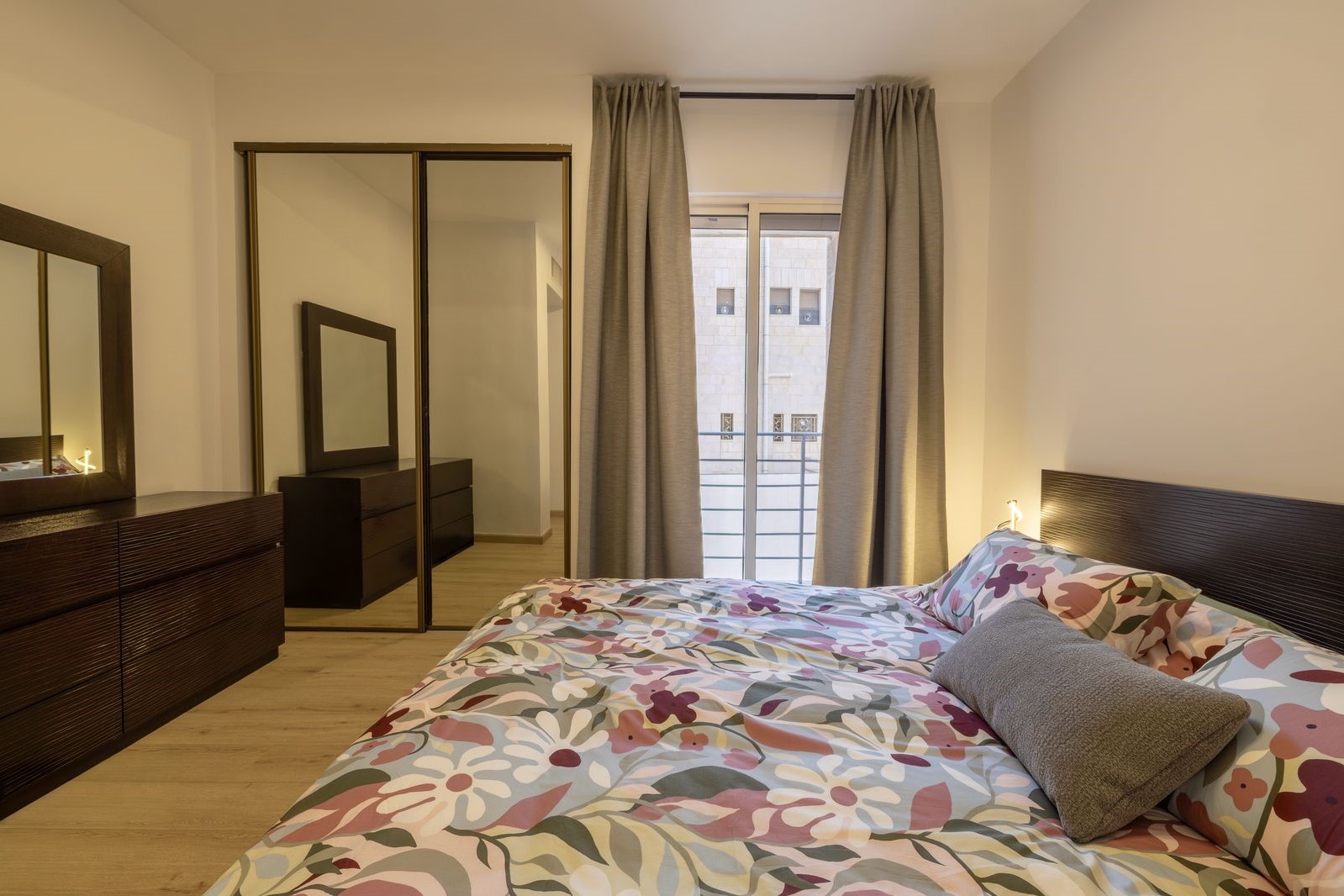 4fd59ac4-e2a3-4704-900d-ef6dbf5934e1 Bedroom with a bed covered in a colorful floral duvet, gray curtains, and dark wood dressers and mirrors around the room.