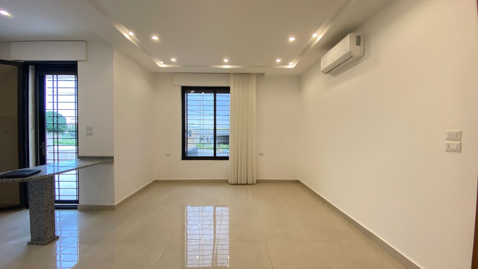 5059b31e-0012-4197-b3a2-bdda3327ccfb Empty living room with glossy tile floor, white walls, recessed ceiling lights, a large window with a white curtain, and a wall-mounted AC.