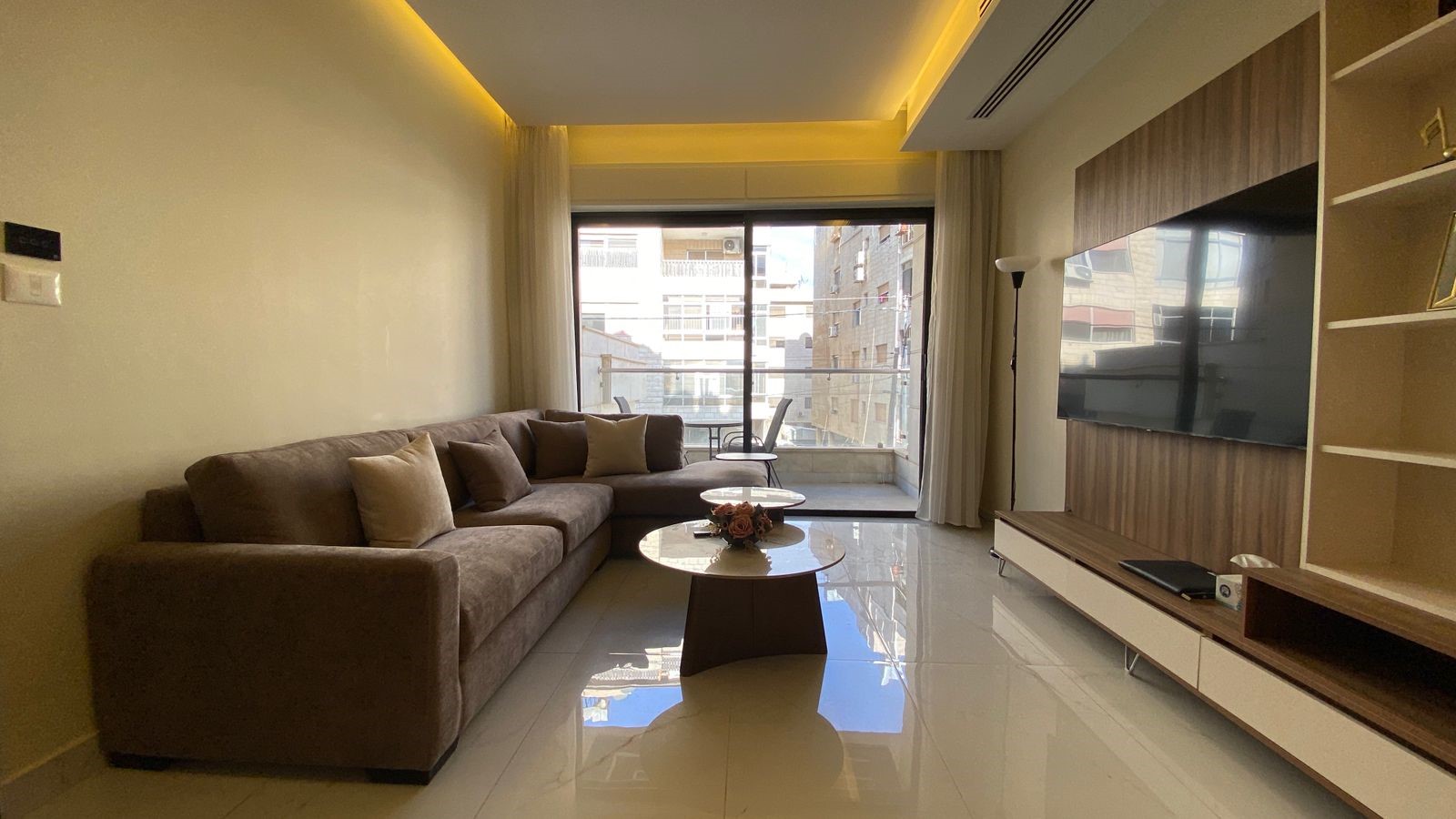 50b9a9bf-f5f9-4678-98b5-e04dd6132fb5 Cozy living room with an L-shaped brown sofa, neutral cushions, and a glass coffee table facing a wall-mounted TV; balcony doors reveal a city view.