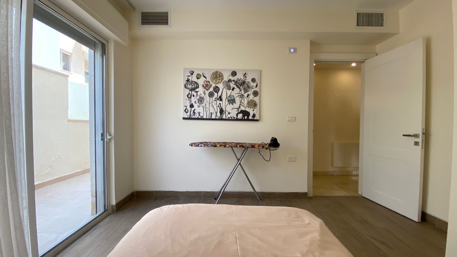 50e140e6-77c6-40cf-ac1e-049efbd8181a Bedroom with a bed in the foreground, ironing board and iron against the wall, and a sliding glass door to the patio on the left.