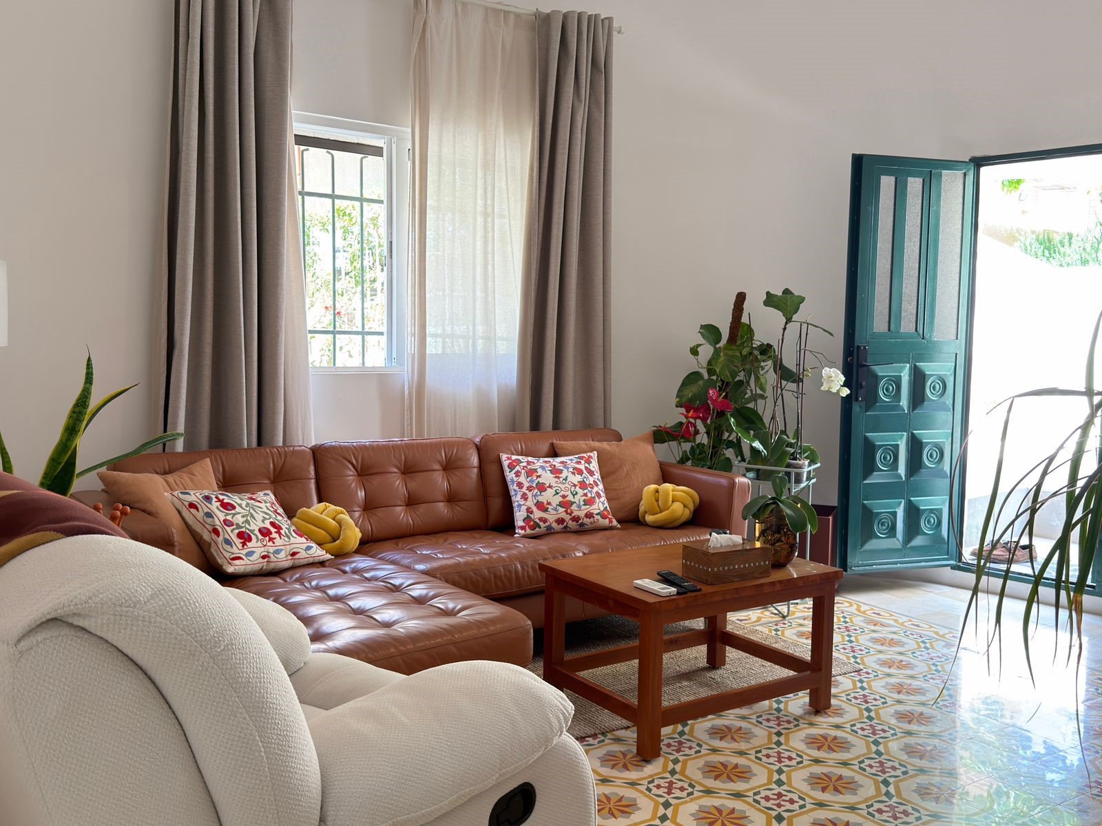 51a82ab3-3311-4c2e-9067-2f00c3187c8d Cozy living room with a tan leather sectional, colorful pillows, a white recliner, and lush green plants by an open teal door.