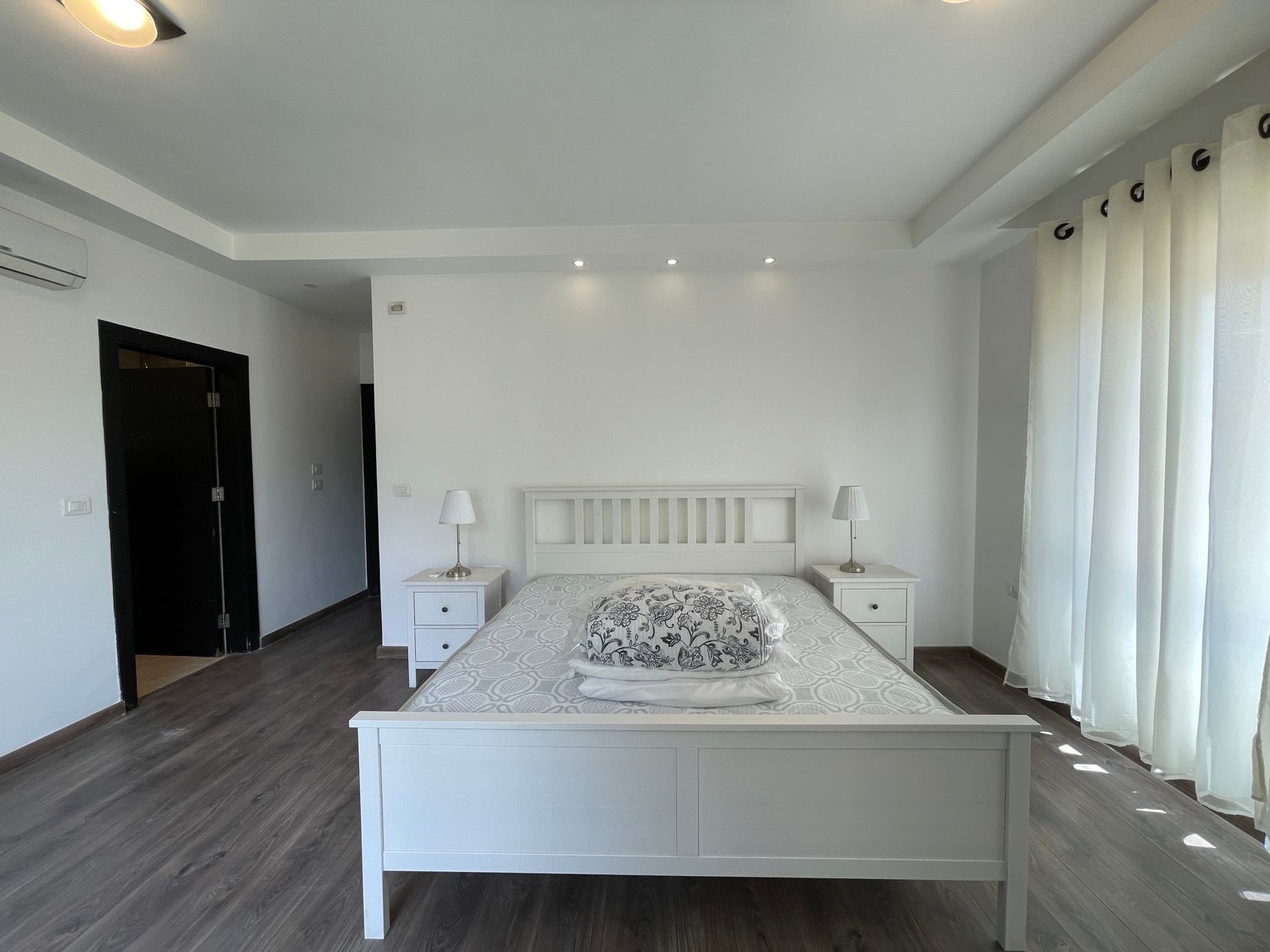 52e8cd21-ca8d-48ff-9c5b-ef5ae336d3e3 Bright white bedroom with a wooden bed frame, two matching nightstands, and soft daylight filtering through large curtains on the right.