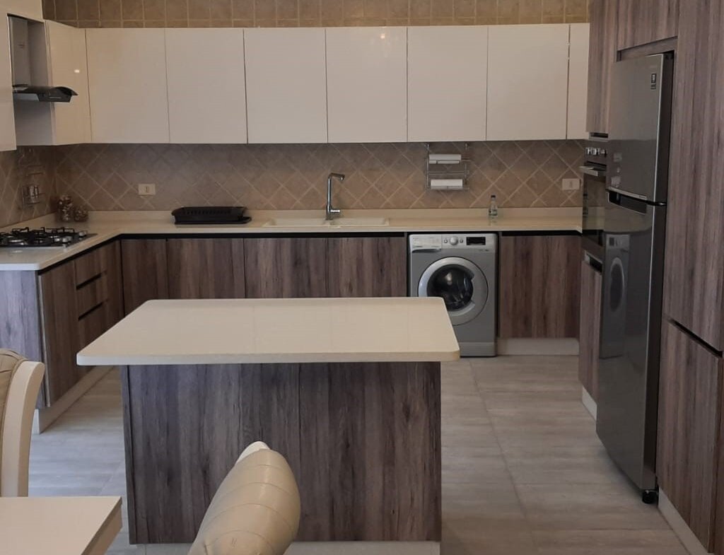 Modern kitchen with white upper cabinets, wood-look lower cabinets, central island, and stainless steel fridge and washer.
