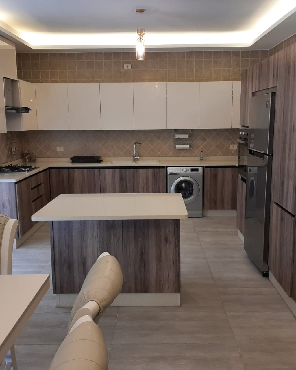 531c8bdf-e8fd-458e-9624-2864afb7b6d5 Modern kitchen with white upper cabinets, wood-look lower cabinets, central island, and stainless steel fridge and washer.