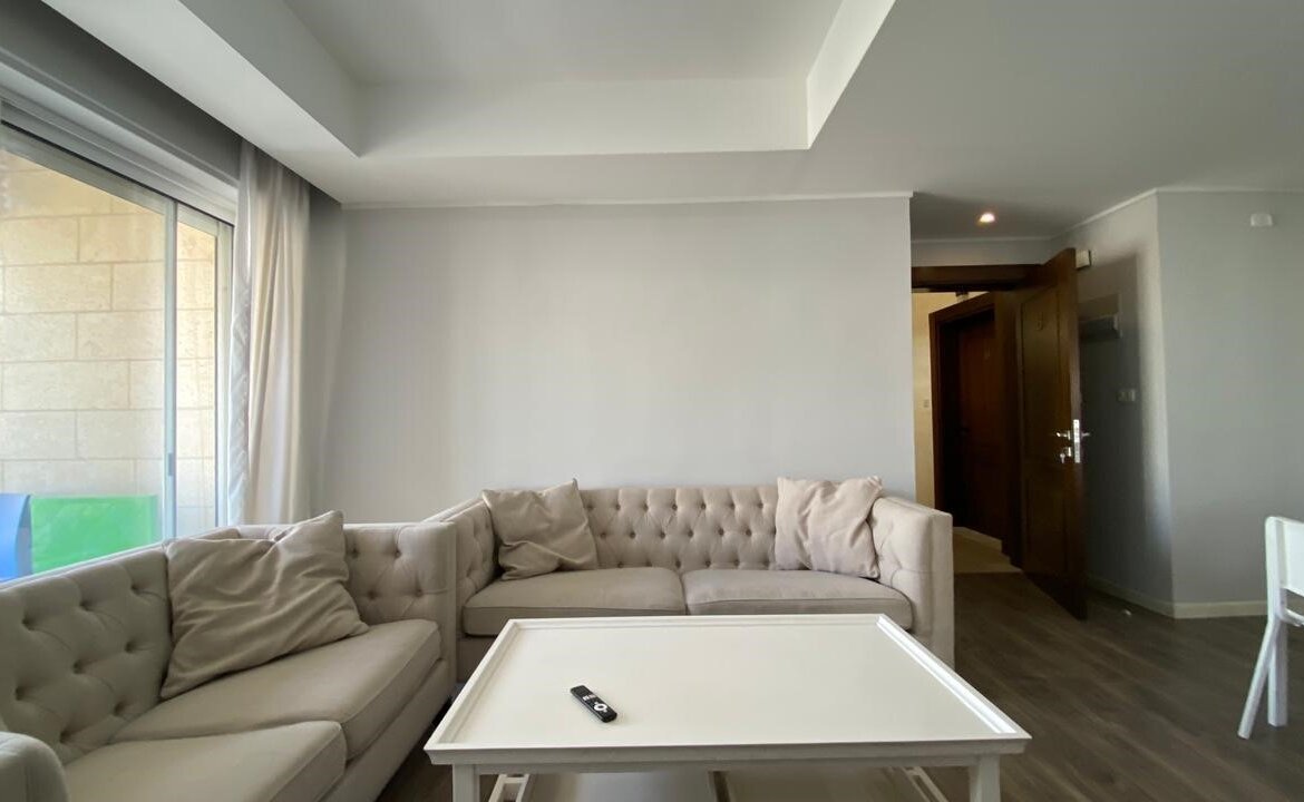 Living room with a pale-taupe tufted sectional, cream cushions, white coffee table, and a remote on it, near a large sliding glass door.