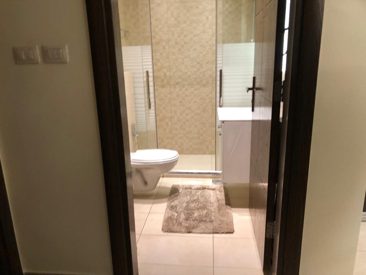 54c70466-b970-4c0c-bc04-0357ad7b753d View into a modern bathroom: toilet at left, glass-enclosed shower in the center, and a white sink with cabinet on the right; beige tiled walls and a gray rug on the floor.