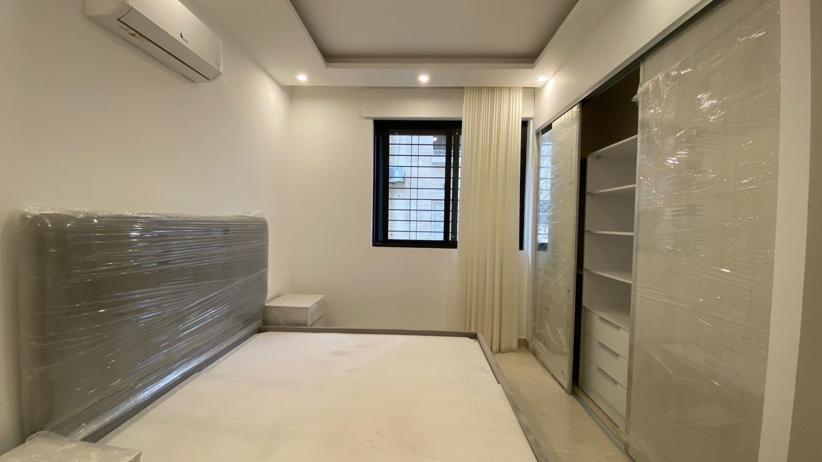55e8c825-7214-4b9b-b33a-aca13cd57f0d Newly furnished bedroom with a bed and a large wrapped headboard, and a built-in wardrobe still covered in plastic.