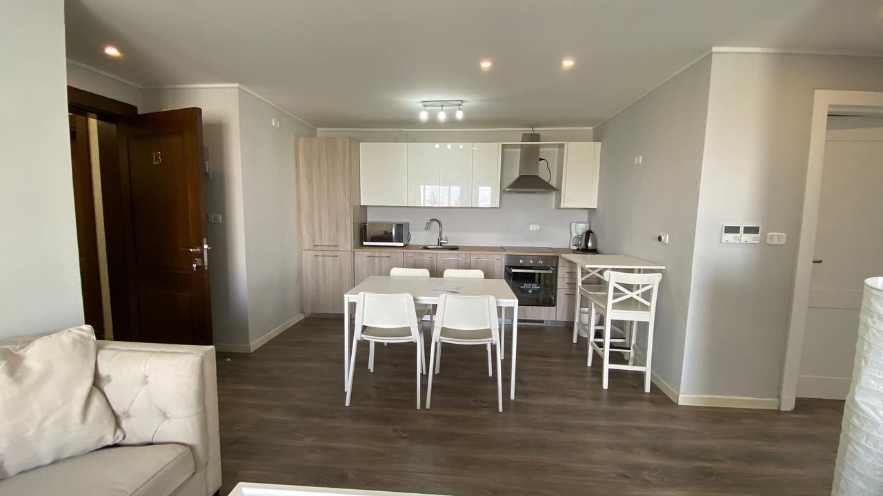 5657f9d7-c065-40f7-842a-0aa45c3fa0e8 Open-plan kitchen and dining area with a white table and four white chairs, wood-tone cabinets, and stainless-steel appliances against light gray walls.