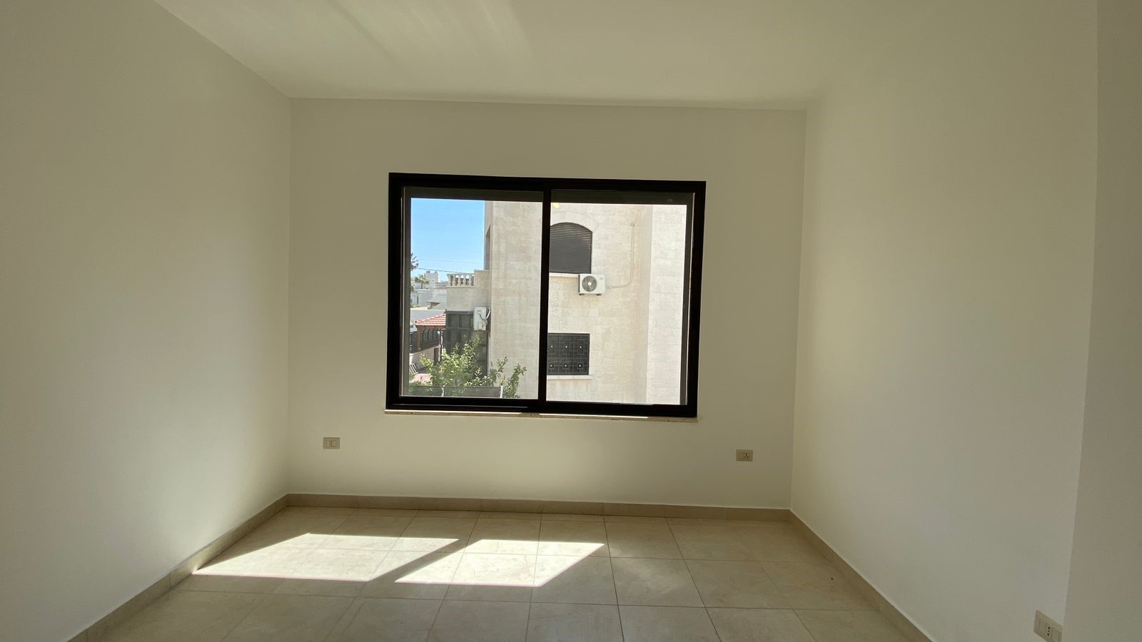 58ac87ce-a810-4522-97bc-6ba8a4b0b56e Empty white room with a large black-framed window, sunlight on tiled floor, view of a neighboring building outside.