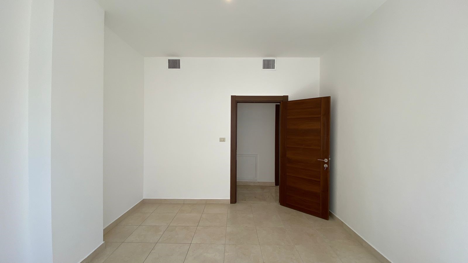 597b22a6-e744-4ea0-aa91-9e13579b0eb9 Empty, well-lit room with white walls, beige tiled floor, and a dark wooden door opened to a small adjacent space; two vents near the ceiling on the back wall.