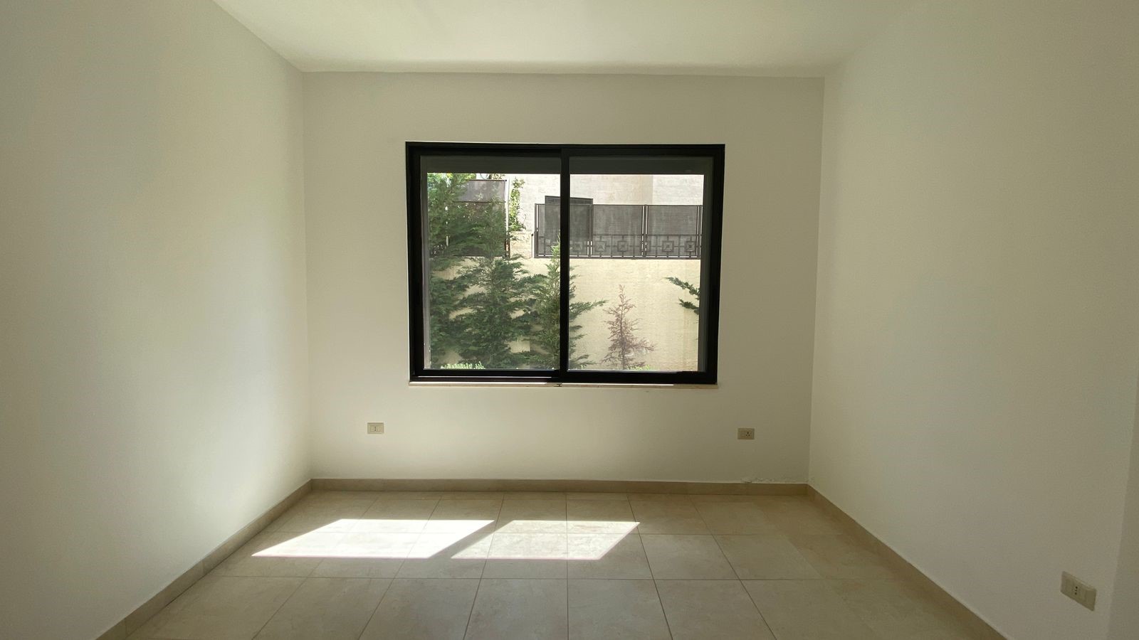 59a04265-33c5-45be-a8c5-8fd2ca16e21a Empty rectangular room with white walls, beige tile floor, and a large window letting in sunlight.