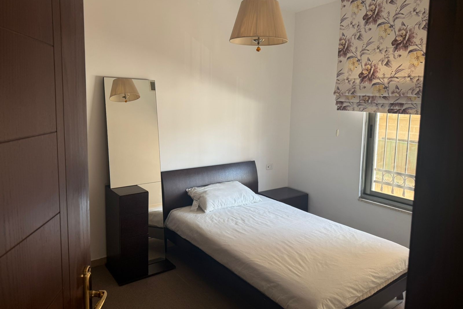 5a5be8e6-281f-4c95-8c4e-3b8517c8d49a Small bedroom with a single bed, white linens, dark wood headboard and matching nightstands, and a floral window shade.