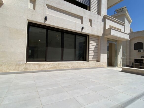 Modern stone building with large glass doors and a wide tiled terrace under bright blue sky