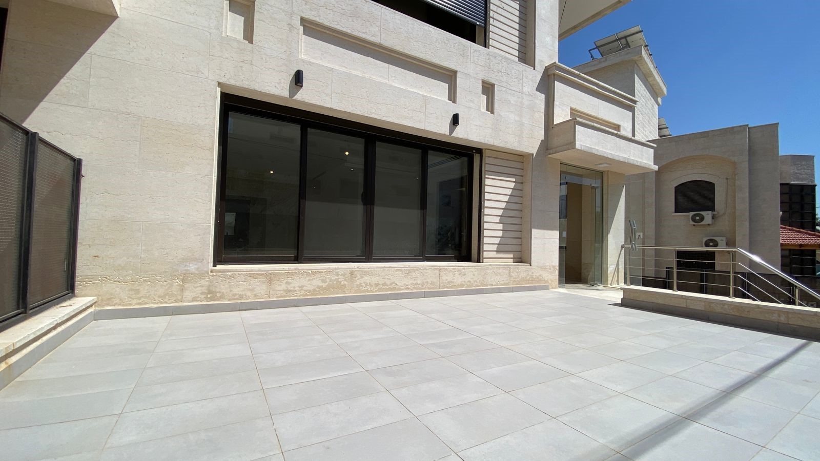 Modern stone building with large glass doors and a wide tiled terrace under bright blue sky
