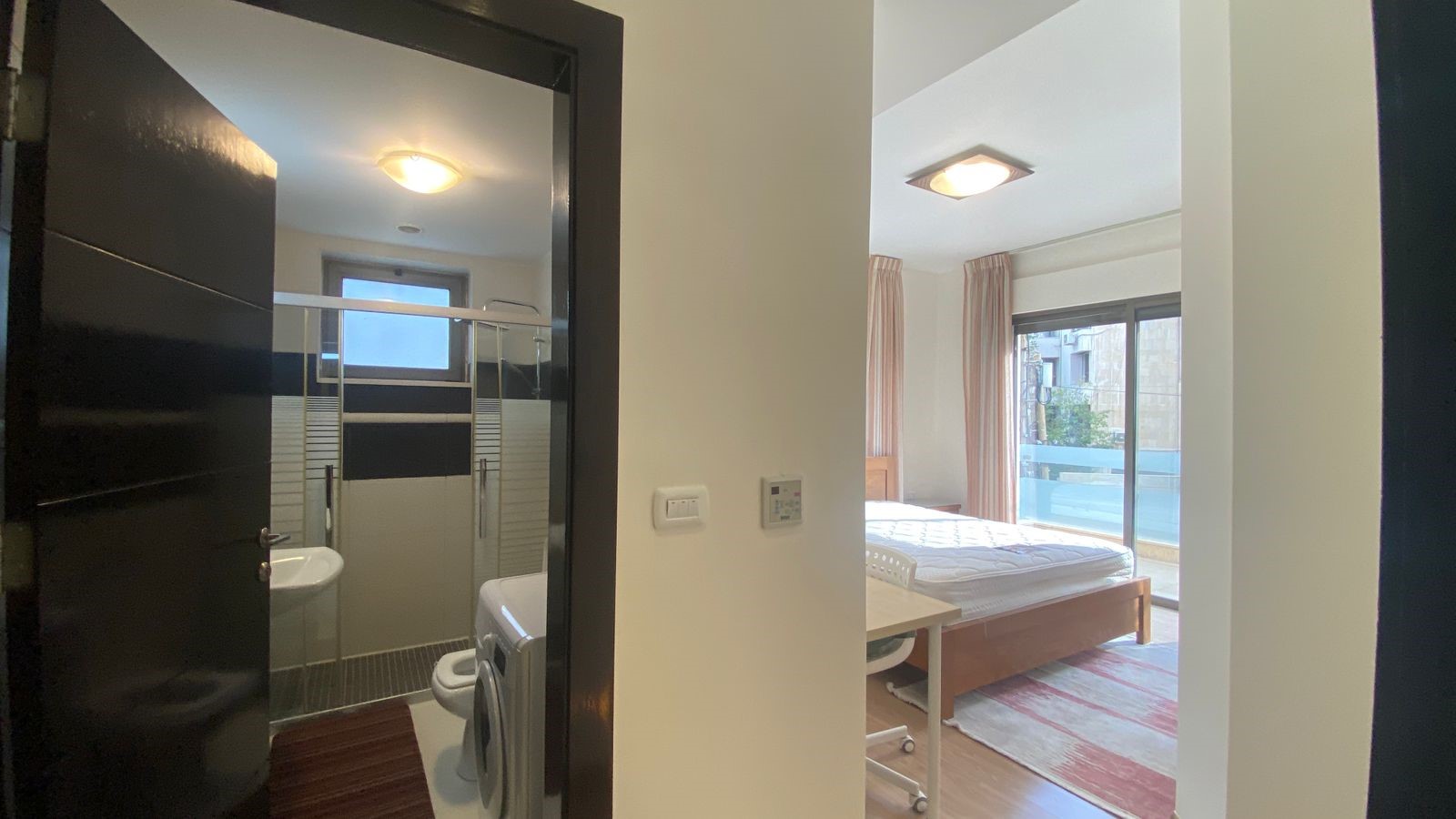 5a95883f-712c-4576-b795-8870fbaa15df Bedroom with a wooden bed, desk, and large window; en-suite bathroom with a shower, toilet, and sink visible on the left.